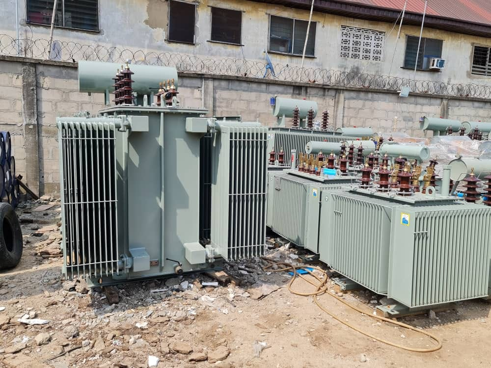 Several oil-immersed distribution transformers with radiator fins, conservator tanks and HV bushings staged in a walled yard